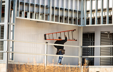 Construction worker carrying large a frame ladder at construction site with steel framing