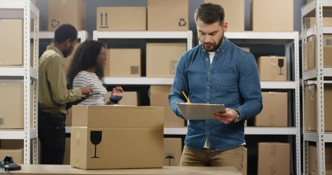 Caucasian postman standing at desk and registering carton parcel in post office store. Mailman writing and filling in invoice. African American male and female postal co-workers standing and talking.