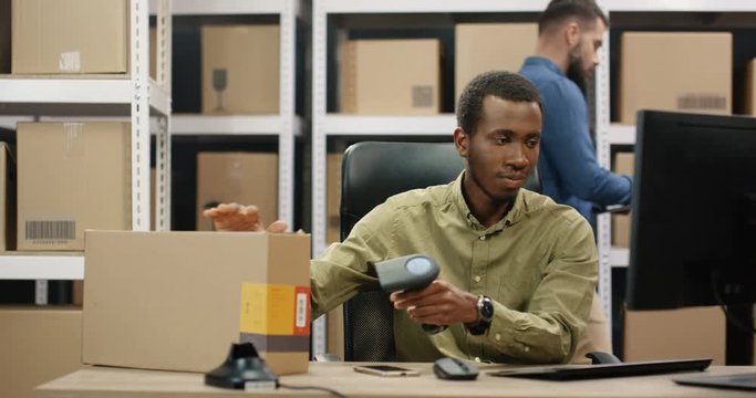 Handsome African American young mailman sitting at table in postal office and working at computer. Male post courier filling in invoice and entering data while scanning bar code of parcel with scanner
