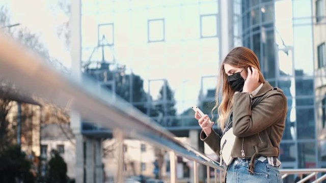 A young lady with a smartphone near a scyscraper is wearing black respirator.