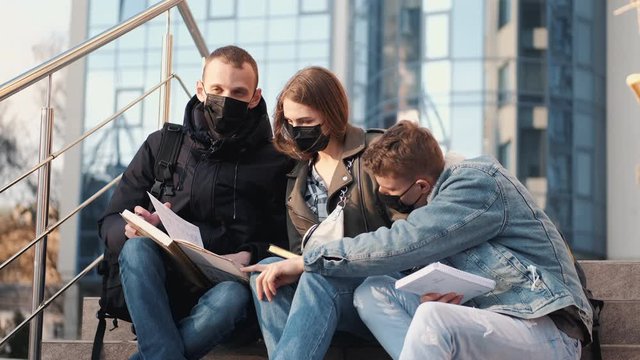 Young people are sitting on stairs outside a bulding wearing black masks.The boys and a girl are reading a book.