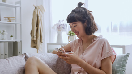 Obraz na płótnie Canvas Attractive smiling asian chinese woman using smart phone while sitting on comfort sofa at home. Communication and coziness concept. charming girl texting message online chatting in couch in morning.