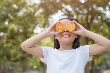 photo Happy Little asian girl child standing showing front teeth with big smile. Covering eyes with orange..fresh healthy green bio background with abstract blurred foliage and bright summer sunlight.
