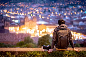 Asian man traveler and photograpaher sitting on viewpoint looking at illuminated cusco city at night. Cusco (Cuzco) is a city in southeastern Peru, near the Urubamba Valley of the Andes mountain range - obrazy, fototapety, plakaty