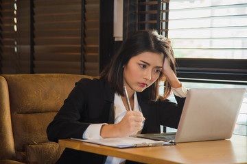 serious young Asian woman in formal suit sitting in front of laptop on wooden table, she is worrying about something in a news in internet that affects business at modern office.