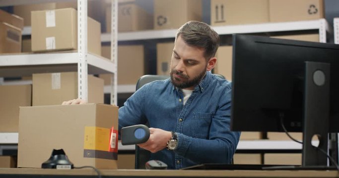 Caucasian handsome mailman working at computer in post office store with parcels. Postman scanning bar code on carton box with scanner, registering it and filling in invoice. Male postal worker.