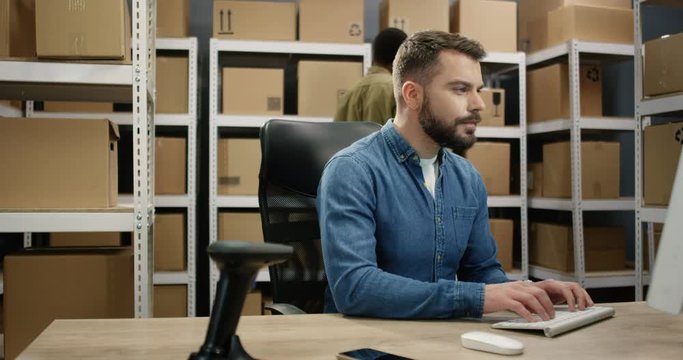 Young attractive Caucasian postman sitting at desk in post office store and working at computer. Mailman typing on keyboard in storage of parcels. Man browsing online.