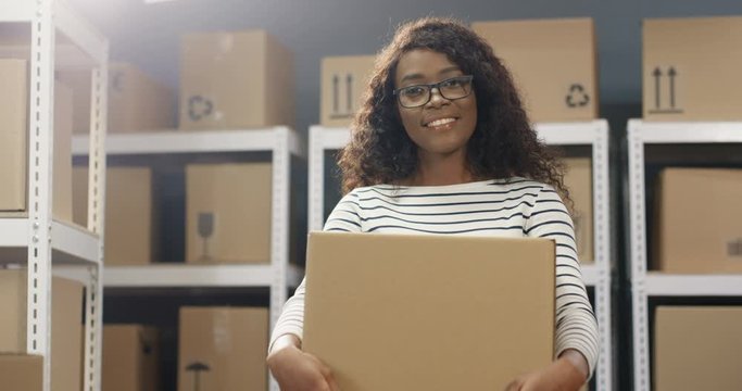 African American happy beautiful woman in glasses smiling cheerfully to camera with carton box in post office store. Postwoman with parcel posing in storage room of parcels. Female courier.