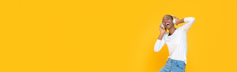 Happy smiling young African American woman dancing while listening to music on headphones isolated on yellow banner background with copy space