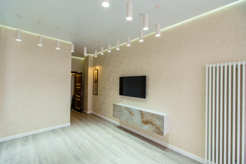 An empty room in beige tones , a TV , a heating battery, and a window in a new apartment with a fresh renovation. There are many hanging cylindrical lamps on the ceiling.