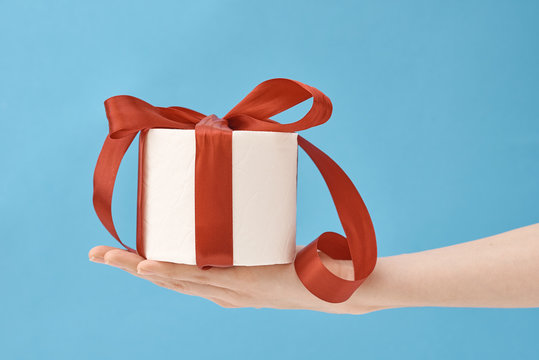 Hand Hold Toilet Paper Wrapped In Festive Ribbon As Gift On Blue Background