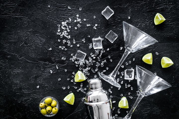 Bar background with martini glasses, shaker, ice, lemon and olives on black table top-down copy space