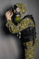 A soldier in camouflage uniform, body armor, in a gas mask with a hood on his head, holds a gun in...