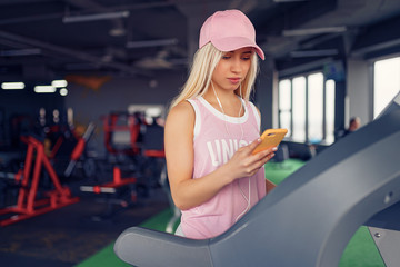 Side view of sporty blonde woman in pink cap prepearing for exercising on treadmill by choosing music on smartphone in gym.