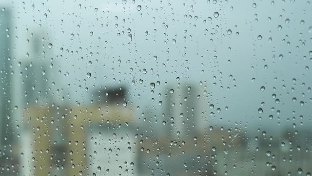 Close up of a glass window on a rainy day with big drops on blurred city ang grey heavy sky background. Stock footage. Transparent water drops on a vertical glass surface.