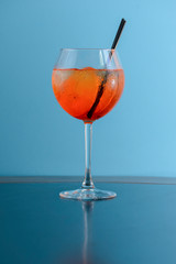 Cold summer alcohol cocktail with ice and orange in a wine glass and black plastic straw on light pastel blue background