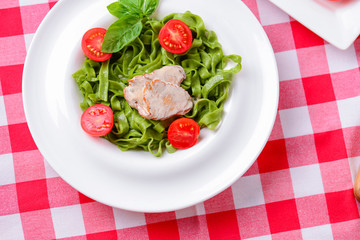 Tagliatelle pasta with pesto, tomatoes and meat stake served on white plate over red plaid tablecloth.