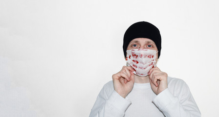 A young man in a white sweater in a hat with a medical mask in blood on a white background. Medicine, coronavirus