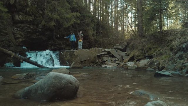 Couple of travelers boyfriend and girlfriend are standing on a rock near a waterfall and taking photos with smartphone. Family enjoying of outdoor traveling. Recreating of life and self isolating.