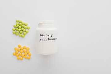 Top view of container with dietary supplements lettering and colorful pills on white background