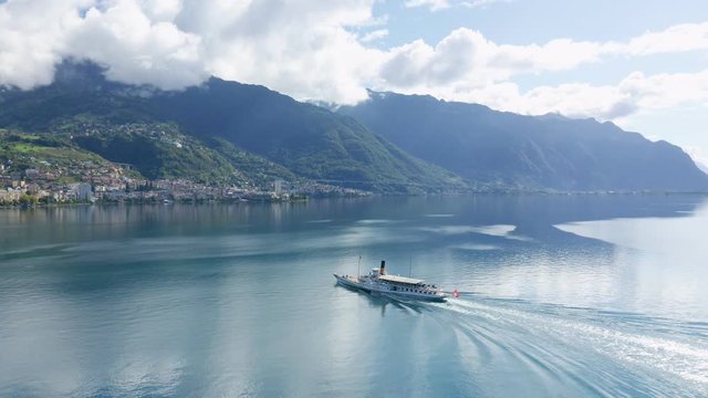 Follow aerial shot of Swiss excursion ship on Lake Geneva. Flag of Switzerland fixed to the stern of boat. Mountains and blue sky with clouds on background