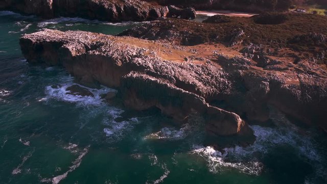 Aerial view of the coast of the Atlantic Ocean during sunrise with warm colors in a cinematographic shot flying from the sea to the coast passing through a natural stone arch over the sea 