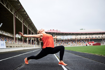 Fototapeta na wymiar Young blond female athlete, wearing orange top and black leggings, doing lunges on a stadium with green grass in summer. Fit sporty woman is doing fitness exercises with motivation to loose weight.
