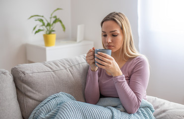 Young attractive woman lying sick on sofa and drinking tea. Sick woman on sofa covered with blanket 