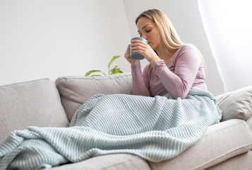 Young attractive woman lying sick on sofa and drinking tea. Sick woman on sofa covered with blanket 