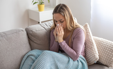 Sick woman blowing nose, sitting under the blanket. Sick woman with seasonal infections, flu, allergy lying in bed. Sick woman covered with a blanket lying in bed with high fever and a flu, resting.