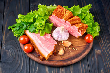 Piece of smoked pork belly with sliced strip and grain bread on a wooden cutting board. Vegetables: tomatoes, lettuce, parsley, garlic and coriander.