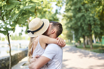 Close-up picture of young couple in love, hugging in park in summer, kissing. Pretty blond girl in stripy overall and straw hat on romantic date with handsome brunette guy in white t-shirt. Meeting.