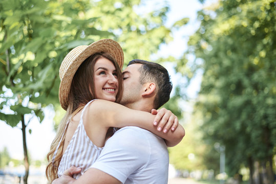 Close-up picture of young couple in love, hugging in park in summer, kissing. Pretty blond girl in stripy overall and straw hat on romantic date with handsome brunette guy in white t-shirt. Meeting.