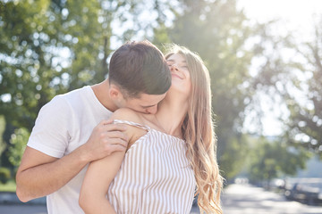 Handsome brunette guy, wearing white t-shirt, hugging and kissing pretty blond woman in stripy overall. Young couple in love, embracing in green park in summer. Romantic relationship. Valentines day.