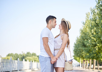 Handsome brunette man, wearing white t-shirt, blue shorts, is hugging pretty blond woman in light straw hat and stripy summer overall. Young couple in love, embracing, looking at each other, smiling.