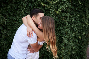 Young couple in love hugging near green bushes trees wall. Pretty blond woman, wearing stripy short overall and brunette man in white t-shirt and blue shorts on romantic date. Romantic relationship