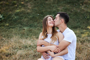 Young couple in love sitting on green yellow grass lawn hugging embracing kissing. Blond woman wearing stripy short overall and brunette man in white t-shirt blue shorts on romantic date. Relationship