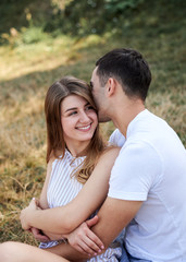 Fototapeta na wymiar Young couple in love sitting on green yellow grass lawn hugging embracing kissing. Blond woman wearing stripy short overall and brunette man in white t-shirt blue shorts on romantic date. Relationship