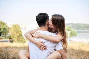 Young couple in love sitting on green yellow grass lawn hugging embracing kissing. Blond woman wearing stripy short overall and brunette man in white t-shirt blue shorts on romantic date. Relationship