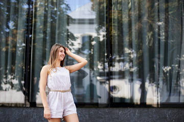 Fototapeta premium Young pretty blond woman, wearing white stripy short overall and sunglasses, standing near big glass building. Three-quarter portrait of slim girl posing for picture in resort. Vacation leisure time