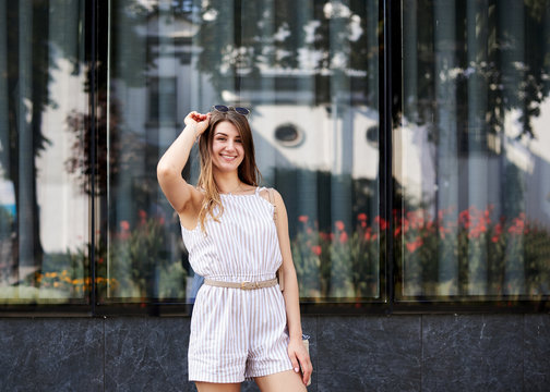Young pretty blond woman, wearing white stripy short overall and sunglasses, standing near big glass building. Three-quarter portrait of slim girl posing for picture in resort. Vacation leisure time
