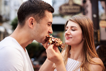 Romantic couple on date eating tasty belgian waffle with chocolate and caramel. Young blond woman, feeding brunette man with yummy desert in summer in the morning. Nutritious breakfast, snack.