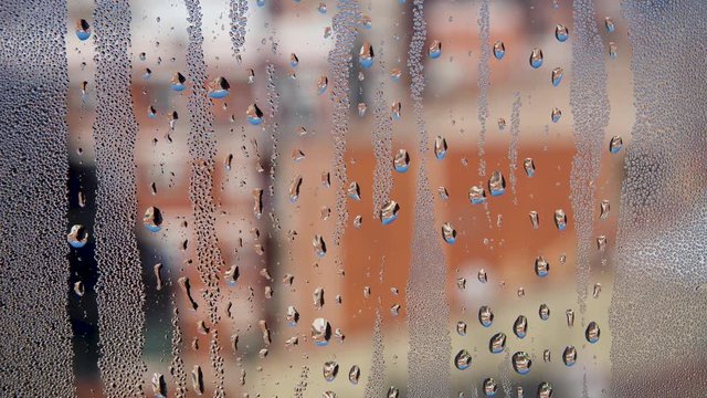 Misted glass of window with water drops and blurred buildings at the background. Water condensate on the glass in a room with high humidity. Camera movement from top to bottom. Water texture.