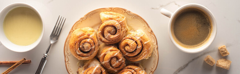 top view of fresh homemade cinnamon rolls on marble surface with cup of coffee, fork and condensed milk, panoramic shot