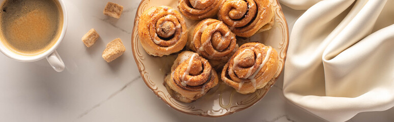 top view of fresh homemade cinnamon rolls on marble surface with cup of coffee, condensed milk and silk cloth, panoramic shot