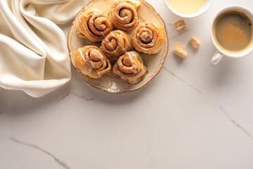 top view of fresh homemade cinnamon rolls on marble surface with cup of coffee, brown sugar, condensed milk and silk cloth