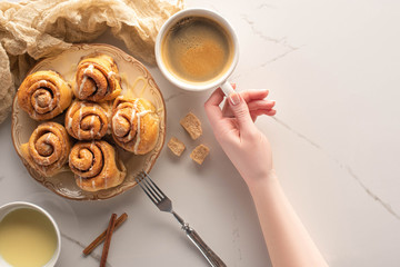 cropped view of woman holding cup of coffee near homemade cinnamon rolls on marble surface with...