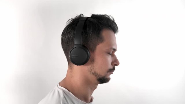 Portrait of caucasian guy with dark hair and beard put on wireless headphones in studio with white background in T-shirt listening music and dancing his hand, half-fase, posing to the camera.