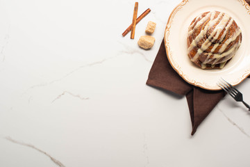 top view of fresh homemade cinnamon roll on plate on marble surface with brown sugar, cinnamon sticks, napkin and fork