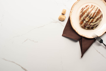 top view of fresh homemade cinnamon roll on plate on marble surface with brown sugar, napkin and fork